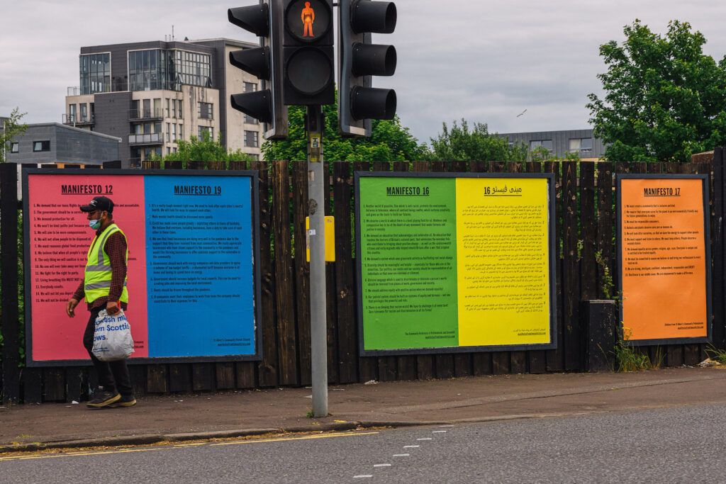 Man in hi-vis vest wearing a face mask walking past 5 largemanifesto posters that are displayed on billboards in Pollokshields Glasgow. The manifestos were made by local communities.